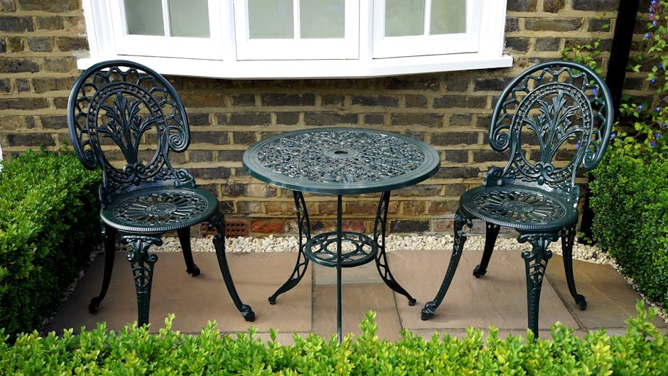 A small outdoor garden seating area featuring two ornate black cast iron chairs with intricate floral and scrollwork designs on the backrests and seats, positioned on either side of a matching round t